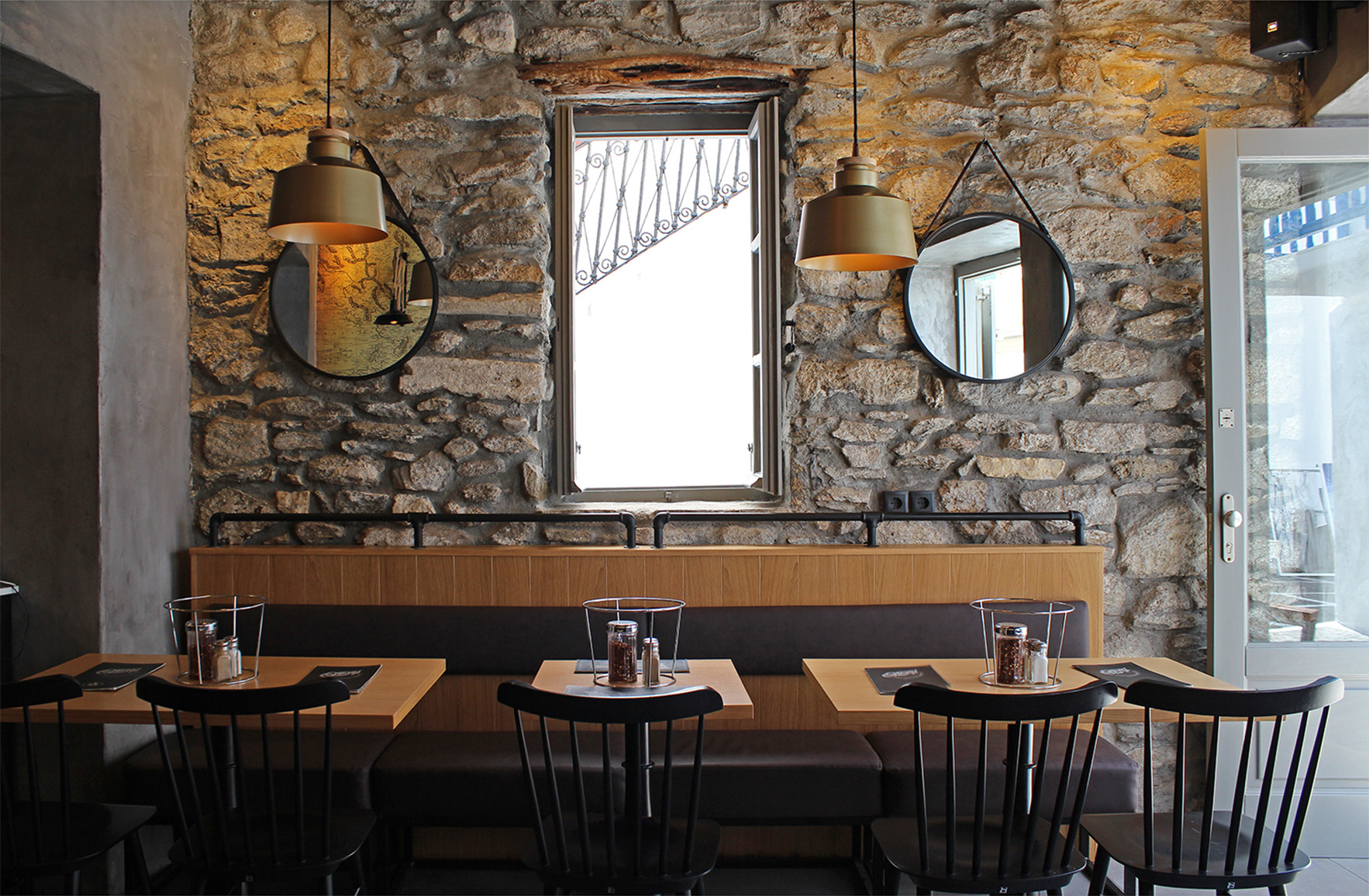 Dining area of L’ Angolo Restaurant in Mykonos, featuring exposed stone walls, wooden tables, black chairs, round mirrors and pendant lighting.