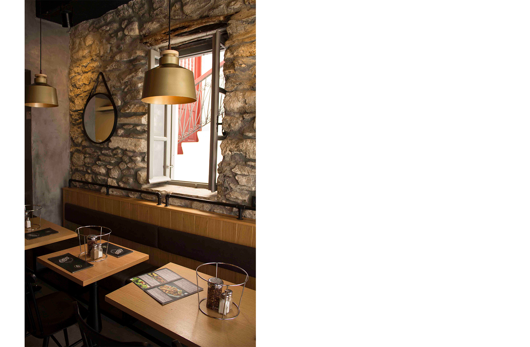 Dining area of L’ Angolo Restaurant in Mykonos, featuring exposed stone walls, wooden tables, black banquette seating, brass pendant lights and an open window.