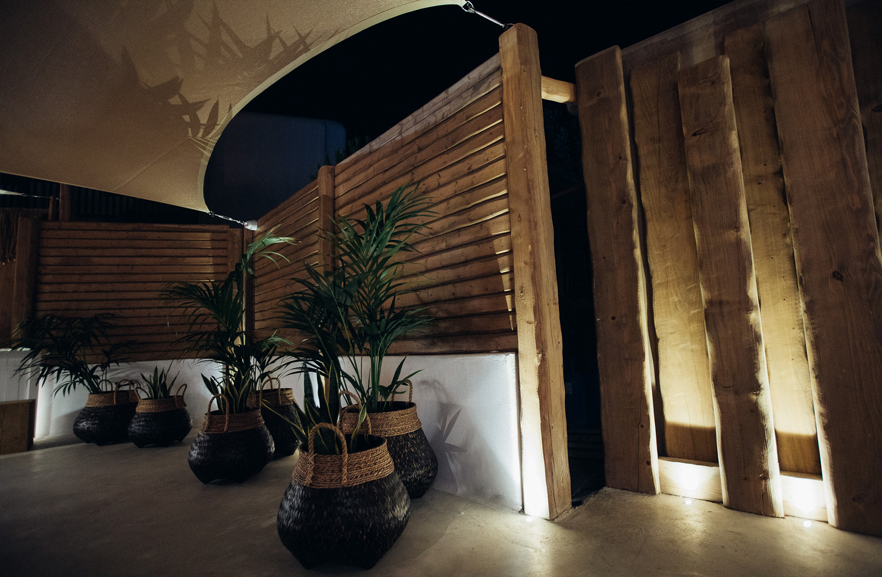 Restaurant in Mykonos featuring timber fencing, shade sails, woven baskets with Mediterranean plants, natural materials, and warm ambient lighting.
