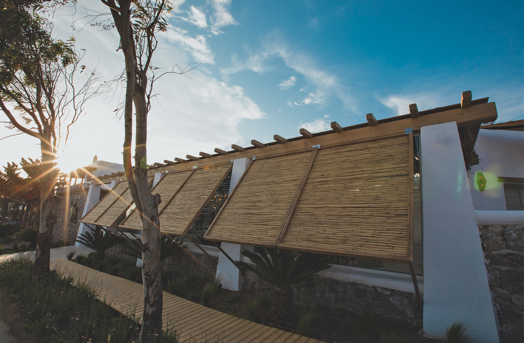 Exterior hospitality architecture in Mykonos with timber pergola, natural shading panels and Mediterranean landscaping by Vicky Basta Designer